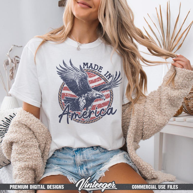May include: White t-shirt featuring a graphic of an eagle with the text "MADE IN America" in a circular design with red and white stripes. The shirt is a casual style.
