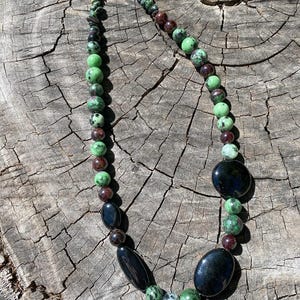 May include: A green, black and red gemstone necklace with black oval-shaped beads. The necklace is on a wooden surface.