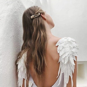 May include: A woman with long brown hair wearing a white top with white lace angel wings on the shoulders.