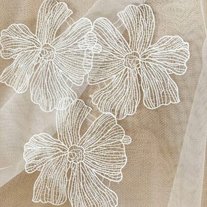 May include: Close-up of white floral lace fabric. Three large, detailed flower appliqués are visible, each with delicate petal patterns. The lace is set against a neutral beige background, creating a soft, elegant aesthetic. Ideal for crafting or apparel.