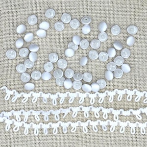 May include: A collection of white pearl-like buttons and white crocheted trim. The buttons are scattered on a beige fabric background.