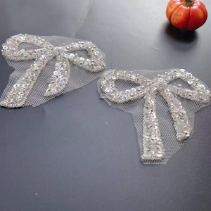 May include: Two silver sequined bow appliques on white mesh. The bows are intricately detailed with small, shiny sequins, creating a sparkling effect. The appliques are likely for embellishing clothing or accessories. A small orange decorative gourd is in the background.