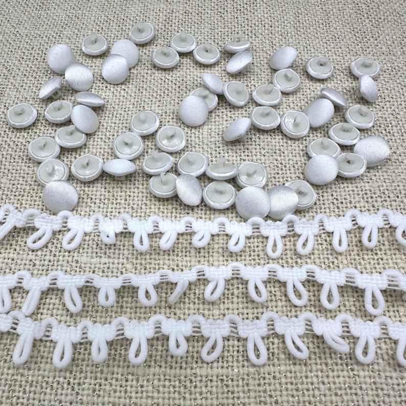 May include: A collection of small, round, white fabric-covered buttons scattered above three horizontal rows of white loop trim. The buttons feature a smooth, satin-like finish, while the trim consists of repeating, interconnected fabric loops.