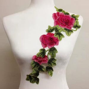 May include: An embroidered floral applique featuring three vibrant pink roses arranged in a diagonal vine pattern, accented with detailed green leaves and stems, designed to be attached to fabric surfaces for decorative embellishment.