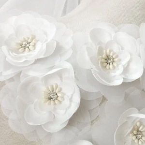 May include: Close-up of white fabric flowers with layered petals and pearl and crystal centers. The flowers are arranged in a cluster, with some overlapping. The fabric appears to be sheer, and the overall effect is delicate and elegant.