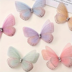 May include: A collection of six sheer butterfly ornaments in pastel shades of blue, pink, purple, peach, and mint green. Each butterfly features delicate translucent wings with subtle spotted patterns and a central silver-toned rhinestone accent.