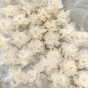 May include: Close-up of ivory-colored fabric flowers with pearl and rhinestone centers. The flowers are arranged densely, creating a textured surface. The petals are delicate and translucent, with a soft, layered appearance. The background is a sheer, light-colored fabric.