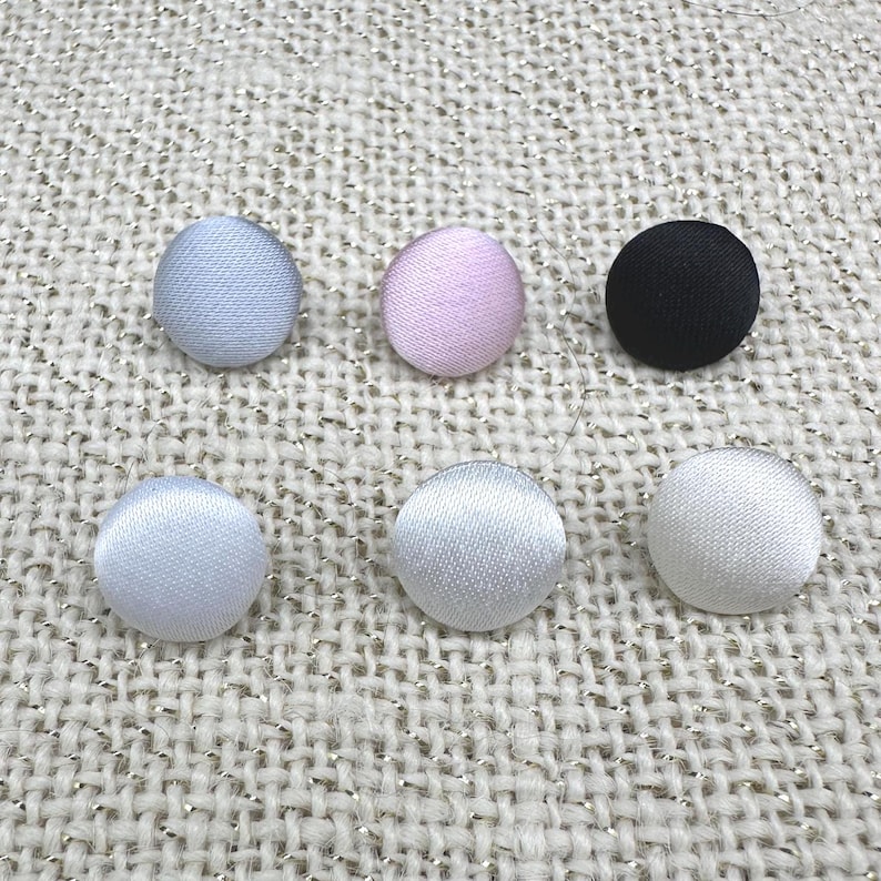 May include: A collection of six round fabric-covered buttons arranged in two rows. The top row features light blue, pale pink, and black buttons. The bottom row consists of three white buttons, all displaying a smooth, woven textile texture.