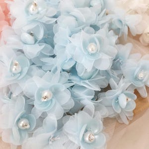 May include: A collection of light blue fabric flowers with delicate petals and pearl-like centers. These handmade flowers are clustered together, showcasing their intricate design and potential for crafting projects or embellishments.