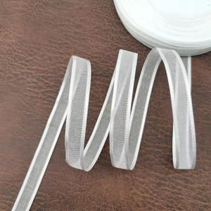 Könnte beinhalten: Weißes, transparentes Band mit Satinrand, in einem wellenförmigen Muster dargestellt. Eine Rolle des gleichen Bandes ist im Hintergrund zu sehen. Geeignet für Bastelarbeiten, Geschenkverpackungen oder zum Hinzufügen einer zarten Note.