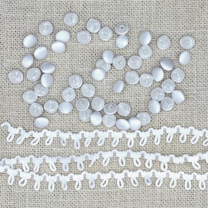 May include: A collection of numerous small, round, white fabric-covered buttons scattered across the frame, accompanied by two long, horizontal strips of white loop trim featuring a repeating, delicate, scalloped edge pattern.