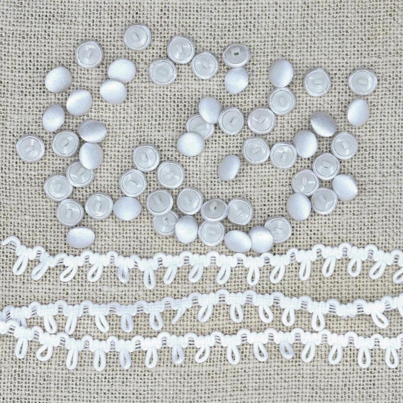 May include: A collection of numerous small, round, white fabric-covered buttons scattered across the frame, accompanied by two long, horizontal strips of white loop trim featuring a repeating, delicate, scalloped edge pattern.