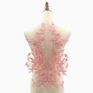 May include: A delicate, light pink floral lace applique. The applique features intricate floral and leaf designs, with a symmetrical pattern. The lace is displayed on a mannequin, showcasing the design's potential for garment embellishment.