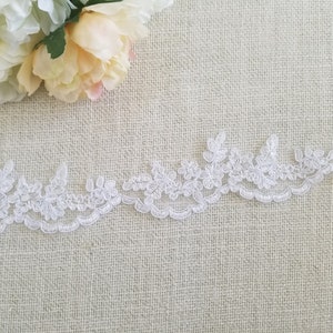 May include: White lace trim with a delicate floral pattern. The lace is shown on a beige fabric background.