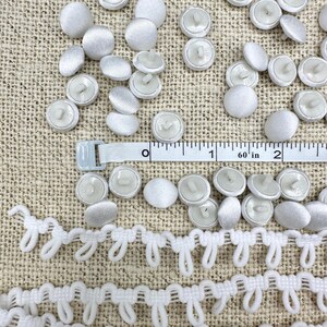 May include: A collection of white fabric-covered buttons and a white loop trim strip. A white measuring tape is positioned across the buttons, showing a measurement of approximately 2 inches in length across the visible section of the tape.