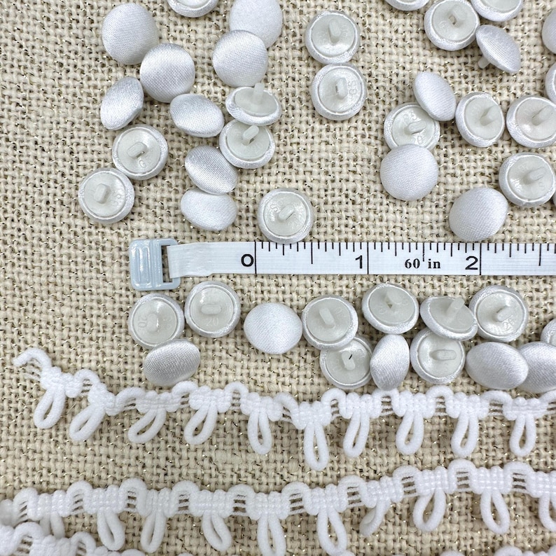 May include: A collection of white fabric-covered buttons and a white loop trim strip. A white measuring tape is positioned across the buttons, showing a measurement of approximately 2 inches in length across the visible section of the tape.