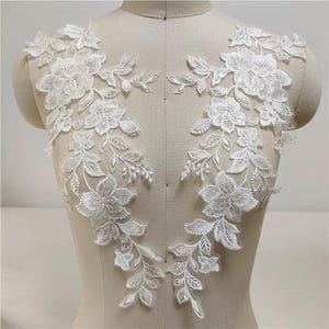 May include: Two white floral lace appliques. Each applique features detailed flower and leaf designs, with delicate textures and patterns. The appliques are displayed on a mannequin, showcasing their potential use in garment decoration.