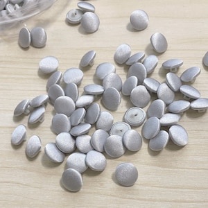 May include: A pile of silver satin buttons, scattered on a light wood surface. The buttons are round and have a smooth, shiny finish.