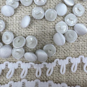 May include: A collection of round, white fabric-covered buttons with plastic shanks on the back, scattered alongside a strip of white decorative loop trim. The buttons feature a smooth, satin-like finish and are arranged in a loose cluster.