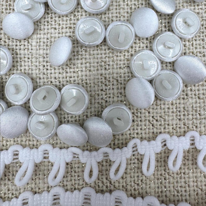 May include: A collection of round, white fabric-covered buttons with plastic shanks on the back, scattered alongside a strip of white decorative loop trim. The buttons feature a smooth, satin-like finish and are arranged in a loose cluster.