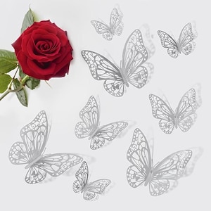 May include: A vibrant red rose with green leaves next to several silver butterfly wall decorations. The butterflies have intricate cut-out designs and are arranged on a white background, creating a delicate and airy aesthetic.