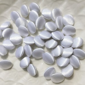 Op de afbeelding: Een verzameling witte satijnen knopen, allemaal dezelfde grootte en vorm. De knopen zijn glad en glanzend en zijn in een willekeurig patroon gerangschikt.