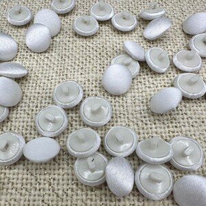 May include: A collection of round, white fabric-covered buttons with a smooth, satin-like finish. Each button features a white plastic shank on the reverse side, designed for sewing attachment. The buttons are scattered across the frame.