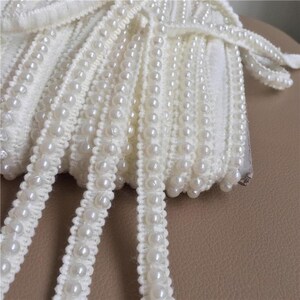 May include: Ivory-colored beaded trim with a delicate, textured appearance. The trim features rows of small, round, pearl-like beads attached to a woven base. The trim is neatly rolled, showcasing its decorative potential for crafts and embellishments.
