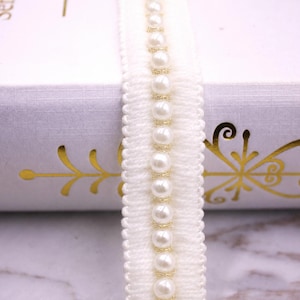 May include: A white fabric trim featuring a central vertical row of round, glossy white faux pearls. The pearls are secured by thin, metallic gold thread accents, and the trim has a textured, scalloped edge on both sides of the pearl strand.