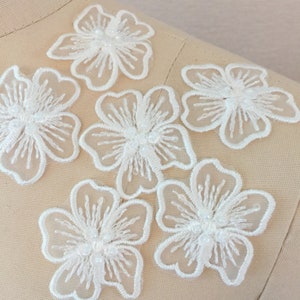 May include: Six white lace flower appliques with pearl embellishments. The appliques are delicate and intricate, perfect for adding a touch of elegance to any project.