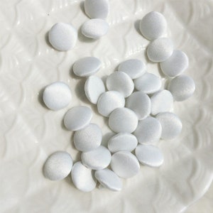 May include: A collection of small, round, white fabric-covered buttons. Each button features a smooth, satin-like finish with a subtle sheen, creating a uniform appearance across the group. The buttons are circular in shape with a slightly domed surface.