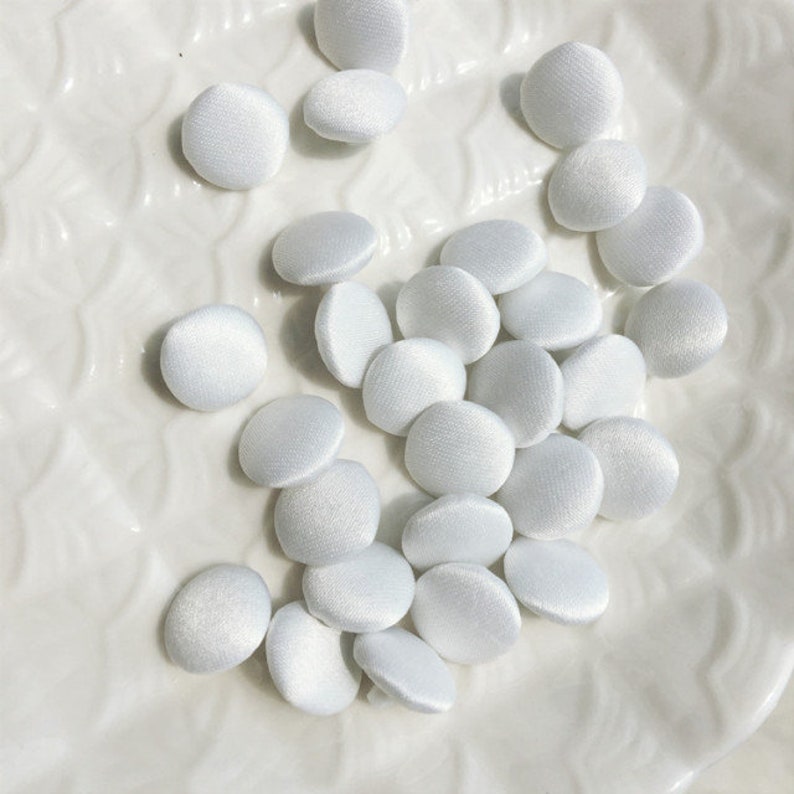 May include: A collection of small, round, white fabric-covered buttons. Each button features a smooth, satin-like finish with a subtle sheen, creating a uniform appearance across the group. The buttons are circular in shape with a slightly domed surface.