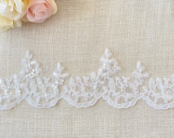 May include: White lace trim with delicate floral designs and small, clear sequins. The trim is shown on a beige burlap background.