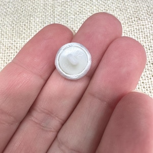 May include: A small, round, white fabric-covered button with a central shank on the back. The back of the button is made of white plastic and features the embossed text 20 and 30, indicating its size and specifications for sewing applications.