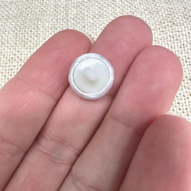 May include: A small, round, white fabric-covered button with a central shank on the back. The back of the button is made of white plastic and features the embossed text 20 and 30, indicating its size and specifications for sewing applications.