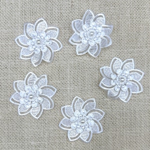 May include: Six white lace flower appliques with pearl centers. The appliques are on a beige fabric background.
