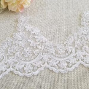 May include: A white embroidered lace trim featuring a scalloped edge with floral motifs and curved decorative lines. The intricate needlework creates a repeating pattern of small blossoms and leaves on a sheer mesh base, forming a continuous border.