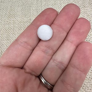 May include: A small, circular, fabric-covered button in a solid white color. The button features a smooth, rounded top surface with a tightly woven textile texture, presenting a clean and uniform appearance suitable for various sewing and craft projects.