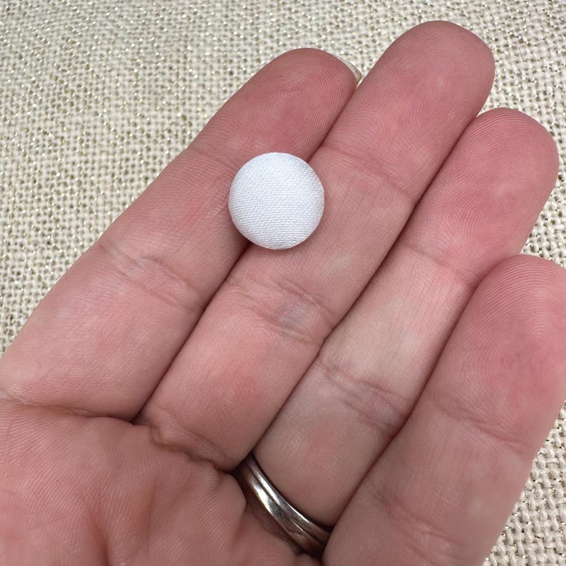 May include: A small, circular, fabric-covered button in a solid white color. The button features a smooth, rounded top surface with a tightly woven textile texture, presenting a clean and uniform appearance suitable for various sewing and craft projects.