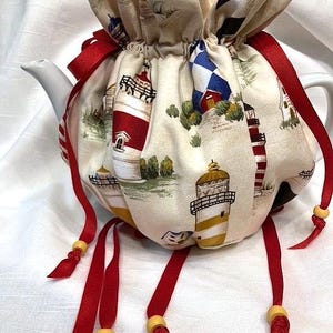 May include: A white ceramic teapot with a fabric cozy featuring a lighthouse print. The cozy is cream-colored with red, white, and blue lighthouses. Red ribbons with wooden beads are tied around the cozy.