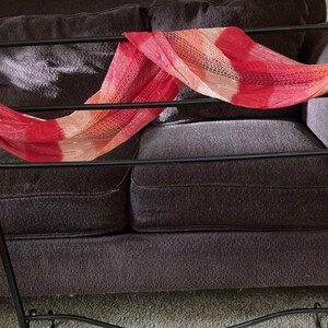 May include: A decorative black metal scarf rack with a red and peach-colored scarf draped across it. The scarf has a striped pattern and fringe on the ends. The rack is in front of a dark brown couch.
