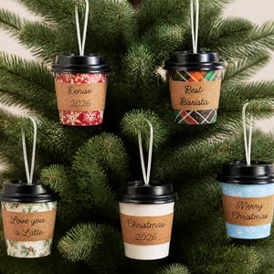 May include: Five miniature coffee cup ornaments with black lids and hanging strings. Each cup features a unique pattern and a brown paper sleeve with text including Denise 2026, Best Barista, Love you a Latte, Christmas 2026, and Merry Christmas.