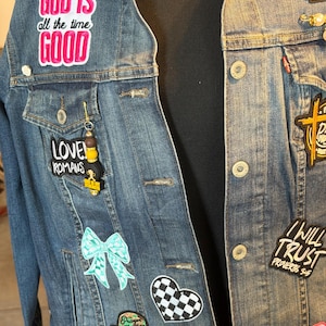 May include: A blue denim jacket adorned with patches and pins. Patches include the phrases "GOD IS all the time GOOD", "LOVE ROMANS", "I WILL TRUST PROVERBS 3:5", and "BLESSED". Additional patches feature a checkered heart, a bow, and a praying woman.