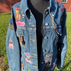 May include: A medium-wash denim jacket adorned with colorful patches and pins. Patches include cartoon figures, text, and brand logos. The jacket is displayed on a black mannequin, with a gold chain detail.