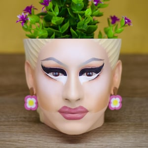 Pode incluir: Um vaso de cerâmica branco em forma de rosto com cabelo loiro, brincos de flores rosa e flores roxas crescendo no topo.