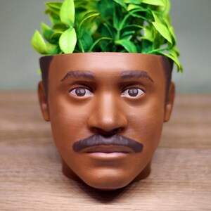 以下が含まれることがあります： 口ひげのある人間の頭の形をした茶色のセラミック製のプランター。プランターには緑色の植物が植えられています。