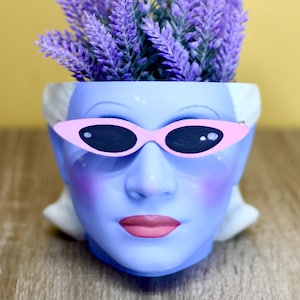 May include: A blue face-shaped planter with pink sunglasses and lavender-coloured faux flowers. The planter has a woman's face with pink blush and lips. The background is a solid yellow colour, and the planter sits on a wooden surface.