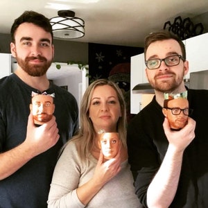 May include: Three people are holding mugs with faces printed on them. The mugs are white with a brown rim. The people are smiling and looking at the camera.