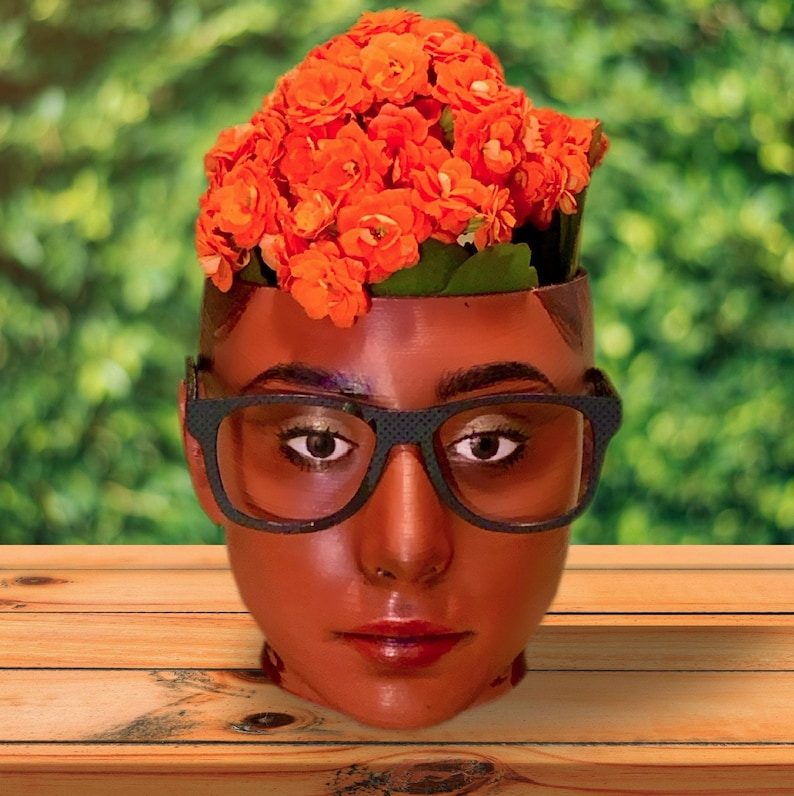 May include: A person's face with orange flowers growing out of the top of their head. The person is wearing black framed glasses.