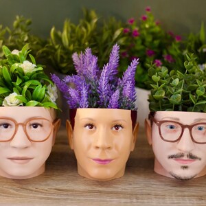 May include: Three ceramic planters shaped like human faces, each with a different plant growing out of the top. The planter on the left has a green plant with white flowers, the middle planter has purple flowers, and the planter on the right has a green plant.